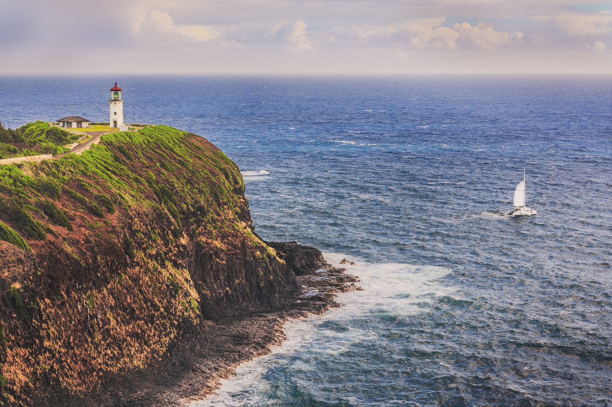 A small white lighthouse on top of a green headland with steep rocky cliffs, waves breaking white below and a sailboat on the open blue ocean.