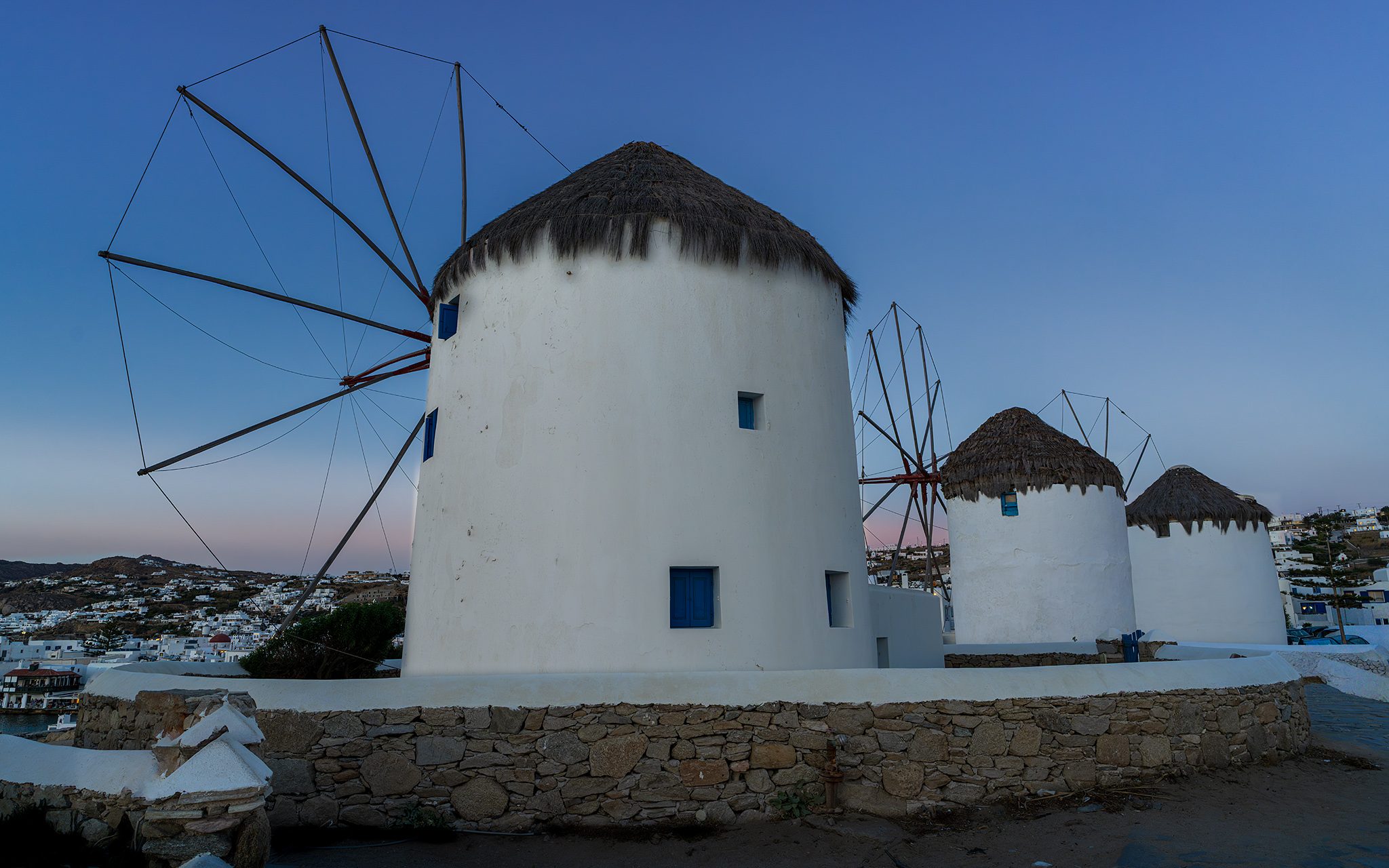 Whitewashed windmills with thatched conical roofs on a low stone wall under a clear blue sky, the nearest mill showing the spokes of its sail wheel.